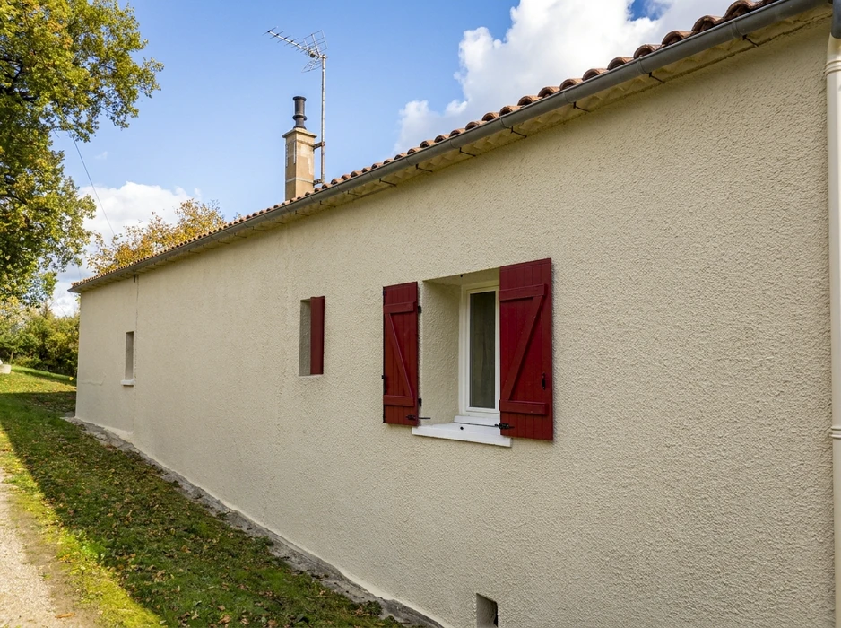Maison de plain-pied style rural français, façade enduit blanc texturé, volets bois peints rouge bordeaux, fenêtre PVC blanc, toiture tuiles canal terre cuite, chéneau zinc, souche de cheminée maçonnée