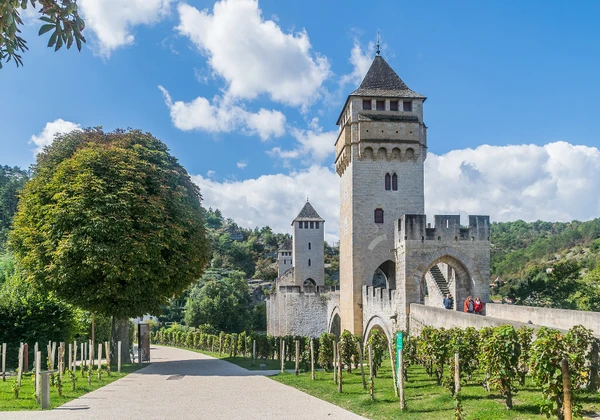 Une photographie réaliste de la ville historique de Cahors dans le Lot-et-Garonne, montrant ses monuments caractéristiques (le Pont Valentré et ses tours emblématiques) surplombant les eaux de la rivière Lot, avec l'architecture médiévale de la vieille ville en arrière-plan, les quais et la végétation environnante. L'image doit capturer l'essence du territoire d'intervention de l'entreprise avec une qualité professionnelle, sans texte ni logo visible.